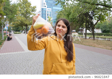 A brunette woman holds a plastic bag of fruit in the park. A brunette woman holds a plastic bag of fruit in the park. 96821847