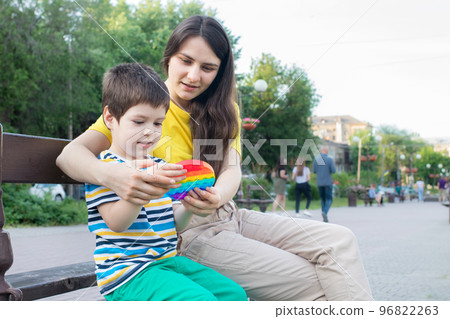 Mom and baby boy 4 years old play with a colored trendy toy Pop it in the park in nature. Antistress sensitive toy or reusable bubble wrap. Trend 2021 96822263