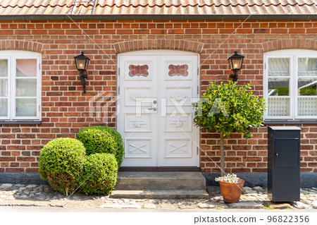 Image of White wooden vintage entrance door with metal side lamps, brick walls and green bushed in front of it 96822356