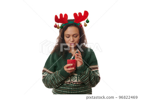 Beautiful dark-haired woman, wearing X-mas patterned warm green sweater and a deer antler hoop, holding a red lit candle, isolated over white background with free advertising space. Merry Christmas 96822469