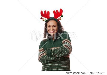Cheerful woman in green Xmas patterned sweater and deer antler hoop, laughing looking at camera, posing with her arms crossed over white background with copy space. Happy atmosphere of winter holidays 96822478