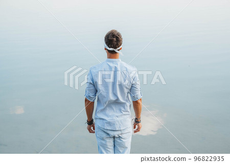 Man handsome dressed in light clothes looks at the water reflex of blue sky. Shoot from the back. Concept of freedom relaxation. Place for text or advertising 96822935