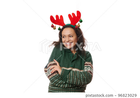 Multi-ethnic joyful attractive woman, wearing deer antler hoop and warm green sweater with Christmas pattern, hugging herself, laughing, looking aside at copy advertising space on white background Multi-ethnic joyful attractive woman, wearing deer antler hoop and warm green sweater with Christmas pattern, hugging herself, laughing, looking aside at copy advertising space on white background 96823029