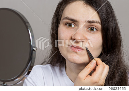 A woman in front of a mirror plucking hairs above her upper lip. Removing the mustache with tweezers yourself. A woman in front of a mirror plucking hairs above her upper lip. Removing the mustache with tweezers yourself. 96823663