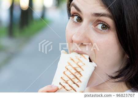 Brunette woman overeats shawarma on a city street. Street fast food pita roll with meat and vegetables. Place for text. 96823707