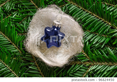 Blue glass star, in a silvery-white angel hair nest, over fresh green fir branches. Star shaped glass bauble to hang on a Christmas tree, placed on metallic tinsel pad. Decorative Christmas ornaments. 96824450