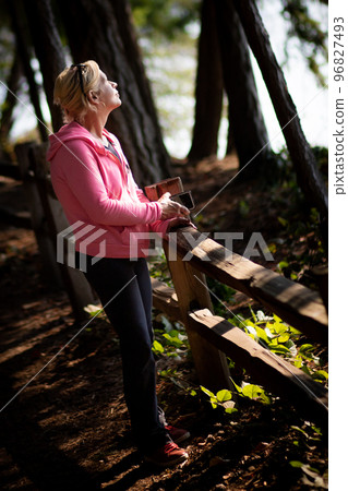 Pretty blonde woman in pink standing next to a fence on a woodland forest trail Pretty blonde woman in pink standing next to a fence on a woodland forest trail 96827493