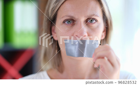 Female with serious face removes adhesive tape from her mouth close up. 96828532