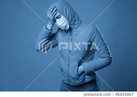 woman wearing hood in a protective face mask and coat over blue background. Model girl in medical mask and spring outfit, studio shot with copy space for your text. Anti-flu, anti-epidemic 96828867