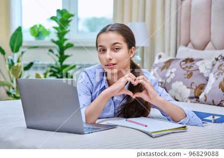 Teenage girl at home with laptop looking at camera showing heart gesture Teenage girl at home with laptop looking at camera showing heart gesture 96829088