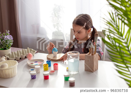 A little girl sits at a white table by the window, paints eggs with a brush and paint in jars. A little girl sits at a white table by the window, paints eggs with a brush and paint in jars. 96830738