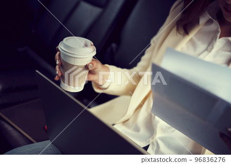 Close-up woman sitting in the back seat of the car with a paperwork in her hand and drinking coffee. 96836276