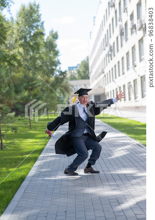 Old happy man in graduation gown jumping outdoors and holding diploma. Vertical. 96838403