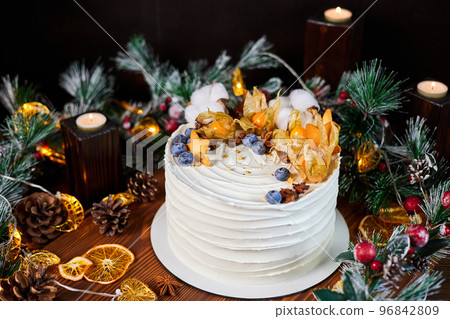 Christmas cake made of chocolate base with cream, decorated with blueberries and physalis against the backdrop of Christmas decor 96842809