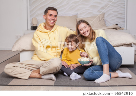Cute laughing smiling parents with child son sitting by the bed and eating popcorn Cute laughing smiling parents with child son sitting by the bed and eating popcorn 96842910