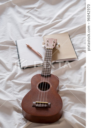 Ukulele guitar and open notebook on white bed. Cozy atmosphere for writing music. 96842970