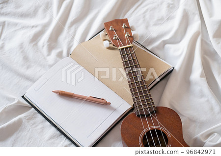 Ukulele guitar and open notebook on white bed. Cozy atmosphere for writing music. Ukulele guitar and open notebook on white bed. Cozy atmosphere for writing music. 96842971