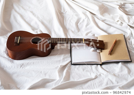 Ukulele guitar and open notebook on white bed. Cozy atmosphere for writing music. 96842974