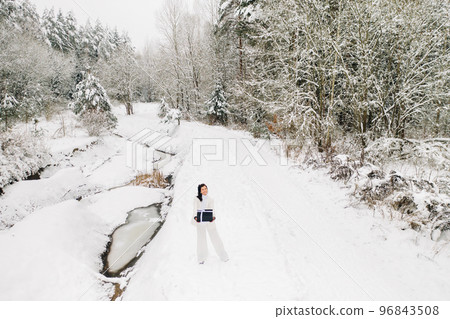 A stylish woman with a white suit with a New Year's gift in her hands in a winter forest. A girl in nature in a snowy forest with a gift box A stylish woman with a white suit with a New Year's gift in her hands in a winter forest. A girl in nature in a snowy forest with a gift box 96843508