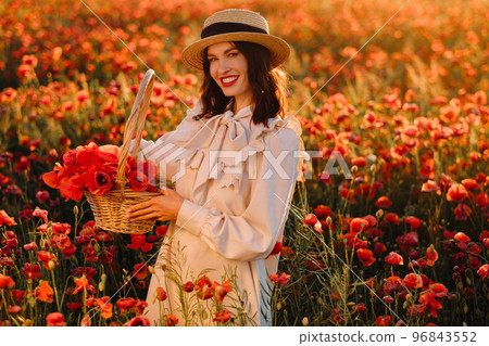 A girl in a white dress and with a basket of poppies is sitting on a poppy field A girl in a white dress and with a basket of poppies is sitting on a poppy field 96843552