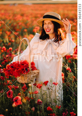 A girl in a white dress and with a basket of poppies walks through a poppy field A girl in a white dress and with a basket of poppies walks through a poppy field 96843553