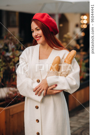 Portrait of a pretty woman in a red beret and a white cardigan with baguettes in her hands strolling through the autumn city Portrait of a pretty woman in a red beret and a white cardigan with baguettes in her hands strolling through the autumn city 96844441