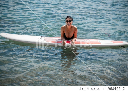 Woman sap sea. Sports girl on a surfboard in the sea on a sunny summer day. In a black bathing suit, he lies on a sap in the sea. Rest on the sea. 96846342