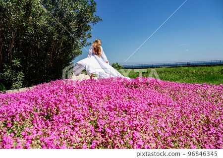 a beautiful woman in a white flowing long dress stay near a beautiful field with pink flowers rear view 96846345