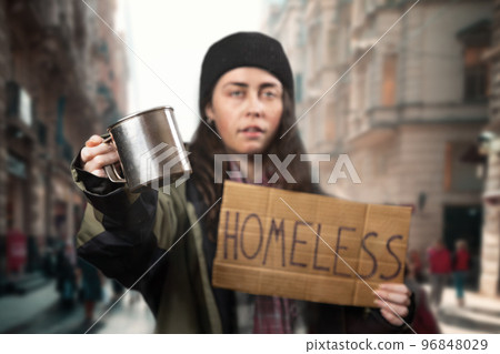 Defocused beggar woman holds a cardboard sign with the text homeless and steel alms cup. Defocused background with street and people. The concept of helping the vagrants Defocused beggar woman holds a cardboard sign with the text homeless and steel alms cup. Defocused background with street and people. The concept of helping the vagrants 96848029