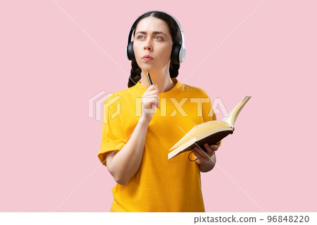 Admission at university. Portrait of thoughtful caucasian young woman wearing headphones holding book and pen at pink background. Concept of online education 96848220
