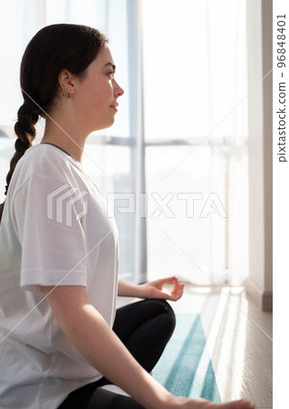 A young pretty woman meditates on a sports mat. A room in the sunlight. Vertical orientation. Side view. The concept of yoga and meditating at home 96848401