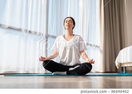 A young pretty woman meditates on a sports mat, closed her eyes. A room in the sunlight. Bottom view. The concept of training at home 96848402