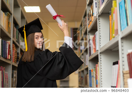 Happy young woman in graduate gown holding diploma in the library.  96848403