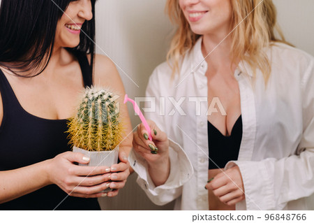 Cactus concept. Unshaven body . One girl is holding a cactus in her hands, and the second girl is shaving a cactus. Symbol of unshaven body parts 96848766