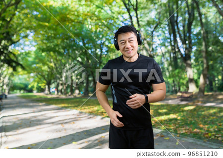 Portrait of a young Asian athlete running in the park, forest, listening to music, podcasts in headphones. He smiles, looks at the camera. 96851210