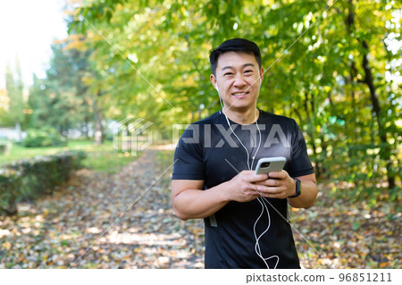 Portrait of a young handsome Asian man who went for a morning jog in the park. He holds a phone in his hand, listens to music in headphones, looks at the camera, smiles 96851211