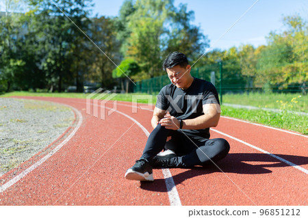 Close-up photo. Sports injury at the stadium. A young Asian man is sitting on a treadmill in black sportswear, holding his leg and knee. He writhes in pain. Medical assistance is required. 96851212