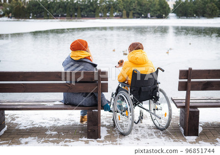 Caucasian woman in a wheelchair and her friend are sitting by the lake with ducks in winter. 96851744