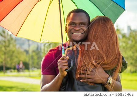 redhaired ginger woman and african american man standing close to each other under umbrella in summer park redhaired ginger woman and african american man standing close to each other under umbrella in summer park 96855462
