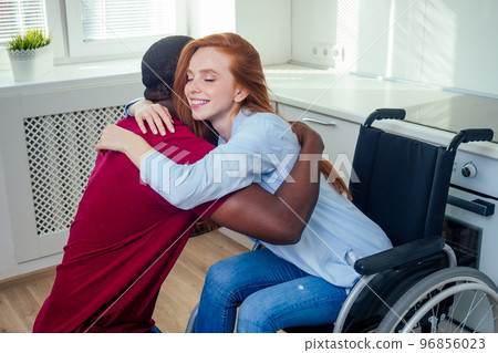 Young bearded handicapped redhaired ginger wife in wheelchair and her african american husband at kichen Young bearded handicapped redhaired ginger wife in wheelchair and her african american husband at kichen 96856023
