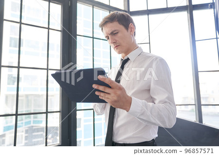 A handsome young businessman using his tablet and typing on it against the background of a window. This person is in the office. With space to copy. High quality photo 96857571