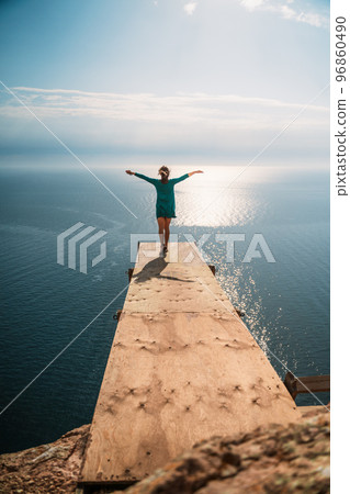 A girl sits on a wooden springboard for jumping with a rope. In a dark green dress with her hands in the air. A girl sits on a wooden springboard for jumping with a rope. In a dark green dress with her hands in the air. 96860490