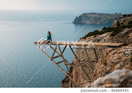 A girl sits on a wooden springboard for jumping with a rope. In a dark green dress with her hands in the air. A girl sits on a wooden springboard for jumping with a rope. In a dark green dress with her hands in the air. 96860491