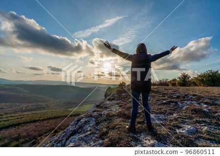 The girl at sunset. The girl stands with her back on the top of the mountain and looks at the sunset, welcomes the sun with her hands up, in Hampi. Meditation, alone with nature, silence. Hands up. 96860511