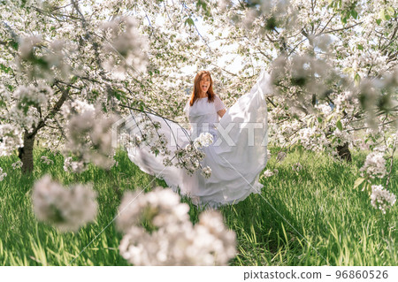 Fantasy woman in long white elegant fashion long dress walks in green spring blossom cherry garden. Happy cheerful girl princess bride. Skirt fabric flies flowing waving in wind motion. Fantasy woman in long white elegant fashion long dress walks in green spring blossom cherry garden. Happy cheerful girl princess bride. Skirt fabric flies flowing waving in wind motion. 96860526