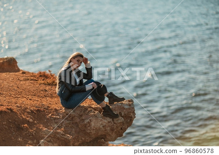 A blonde girl in a stylish black leather jacket is sitting on the seashore. A blonde girl in a stylish black leather jacket is sitting on the seashore. 96860578