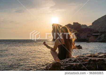 Woman tourist enjoying the sunset over the sea mountain landscape. Sits outdoors on a rock above the sea. She is wearing jeans and a blue hoodie. Healthy lifestyle, harmony and meditation Woman tourist enjoying the sunset over the sea mountain landscape. Sits outdoors on a rock above the sea. She is wearing jeans and a blue hoodie. Healthy lifestyle, harmony and meditation 96860865