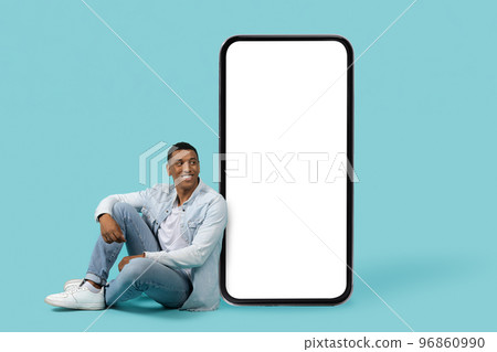 Smiling young african american guy in casual sit on floor, look at huge smartphone with empty screen Smiling young african american guy in casual sit on floor, look at huge smartphone with empty screen 96860990