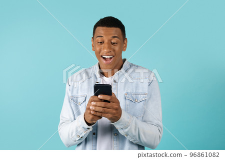 Surprised smiling young african american guy in casual typing on smartphone isolated on blue background 96861082