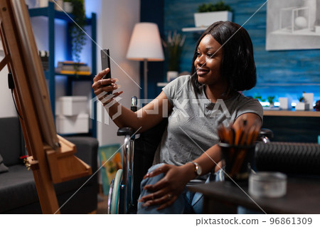 Young female artist sharing finished sketch masterpiece on social media via smartphone from art studio at home. African american professional cartoonist wheelchair user showing artwork via video call. 96861309